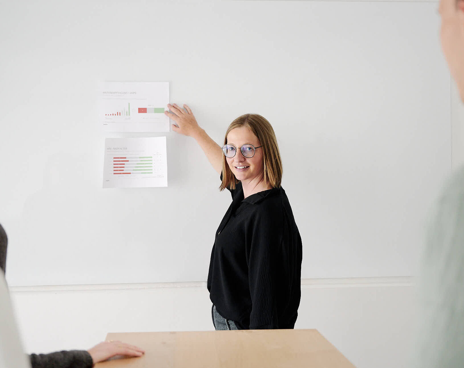 Lächelnde Frau mit Brille zeigt bei einer Präsentation auf Grafiken an der Wand.