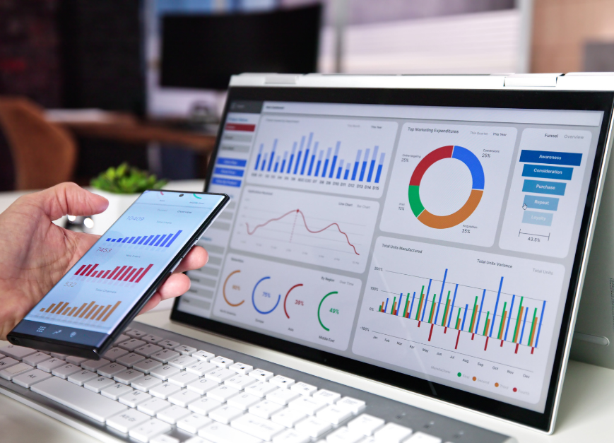 Eine Hand hält ein Smartphone mit Diagrammen vor einem Laptop mit detaillierten Datenanalysen.