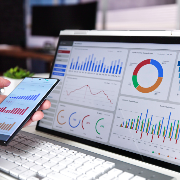 Eine Hand hält ein Smartphone mit Datenanalysen vor einem Laptop mit Business-Dashboards.