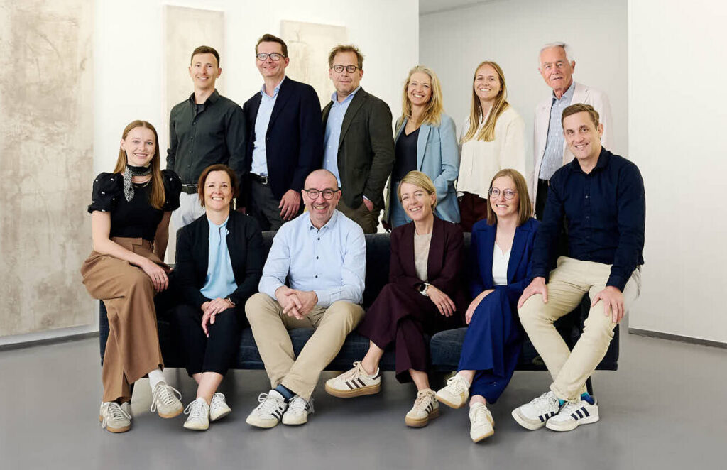 Ein lächelndes, professionell gekleidetes Team posiert gemeinsam in einem hellen, modernen Raum.