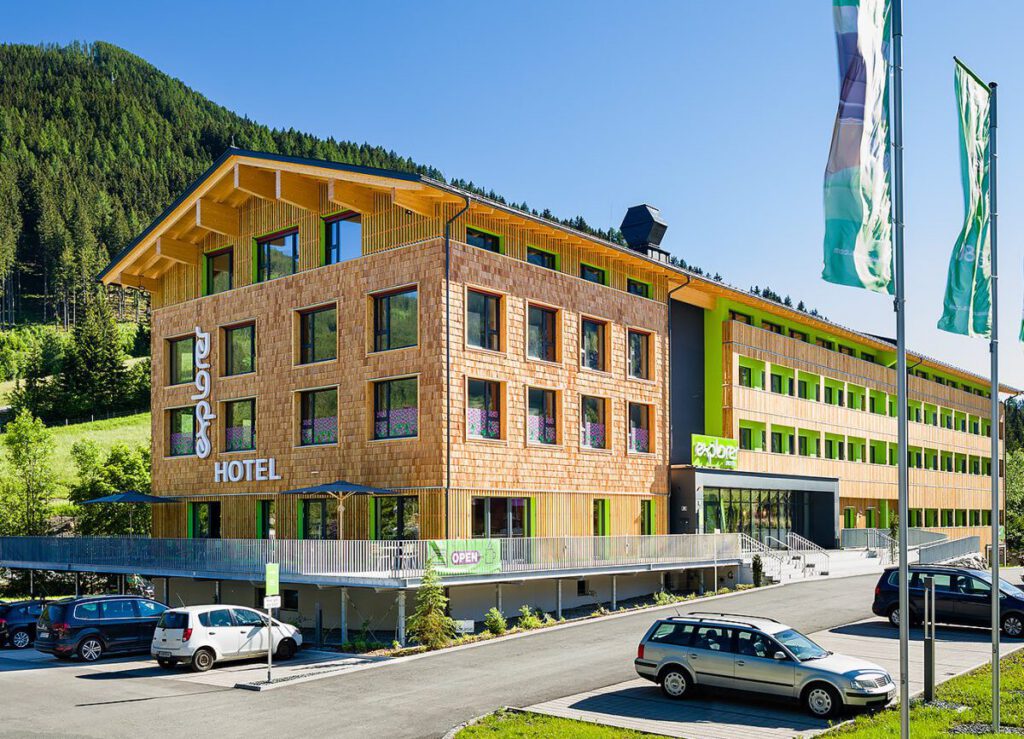 Modernes Explorer Hotel mit Holzfassade und grünen Akzenten vor einer bewaldeten Bergkulisse.