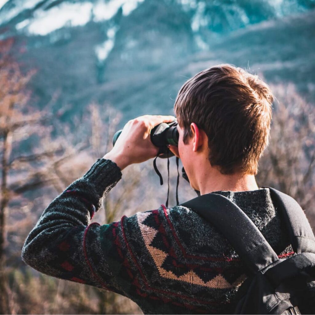 Ein Wanderer mit Rucksack blickt durch ein Fernglas in die Berge.