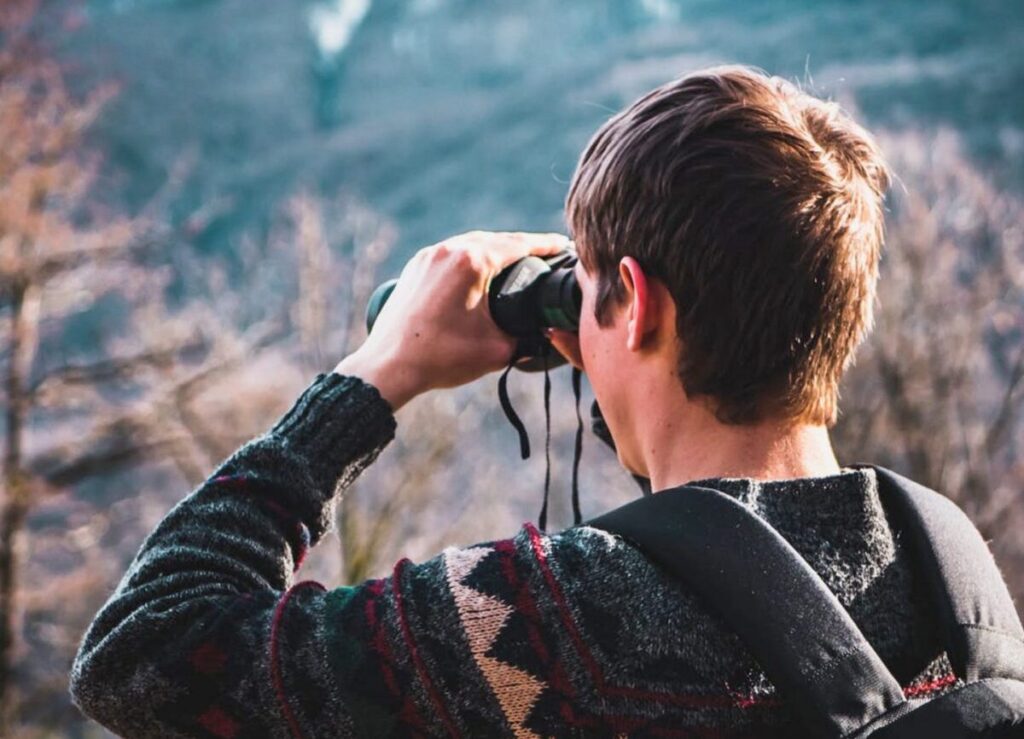 Ein junger Wanderer blickt durch ein Fernglas in eine bergige Naturlandschaft.