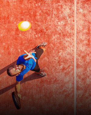 Padel-Spieler im blauen T-Shirt schlägt aus der Vogelperspektive einen Ball auf rotem Platz.