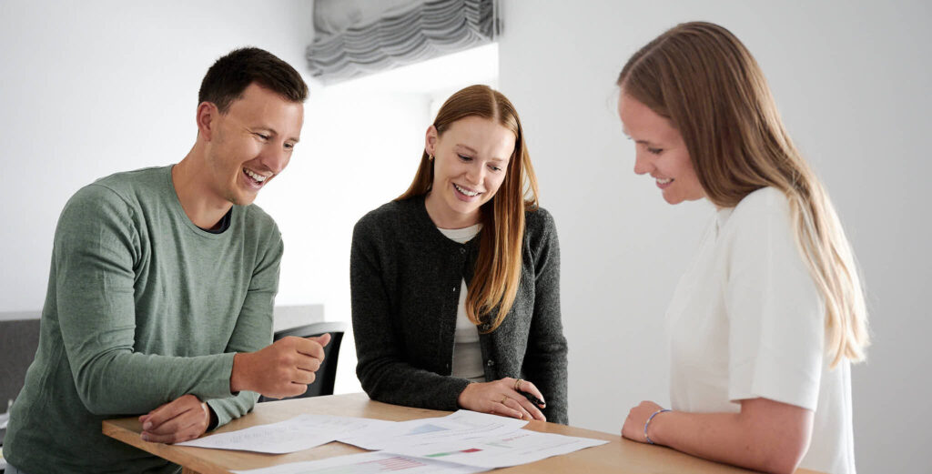 Drei lächelnde Personen sitzen an einem Tisch und besprechen gemeinsam Dokumente.