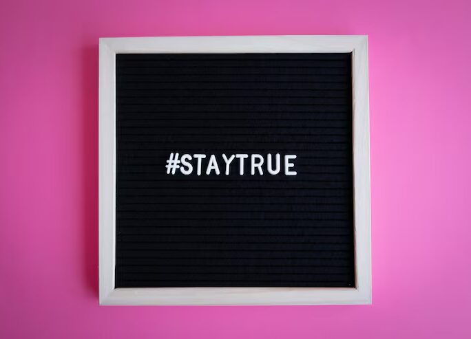 Schwarze Buchstabentafel mit weißem Hashtag STAYTRUE vor einem pinken Hintergrund.