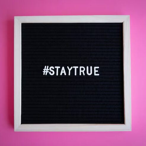 Schwarzes Steckbrett mit weißem Schriftzug Hashtag STAYTRUE auf einem leuchtend pinken Hintergrund.