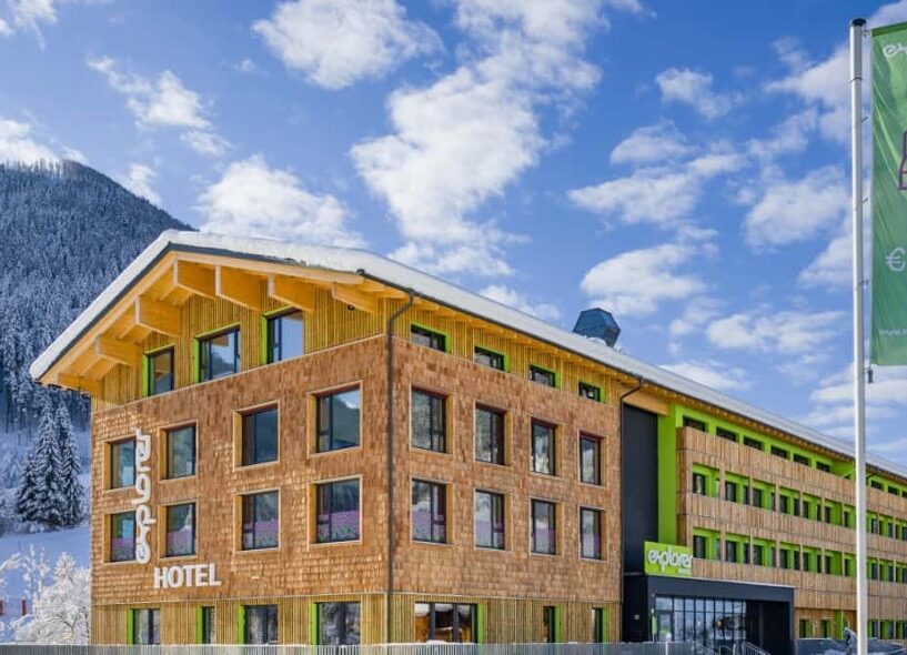 Modernes Explorer Hotel aus Holz vor verschneiter Bergkulisse unter blauem Himmel.