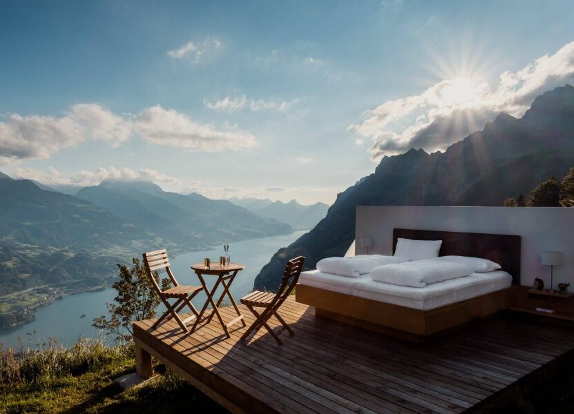 Freiluftbett auf einer Holzterrasse mit Panoramablick auf einen See und sonnige Berge.