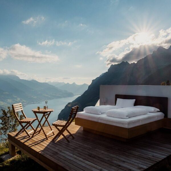 Doppelbett auf einer sonnigen Holzterrasse mit Panoramablick auf Berge und einen See.