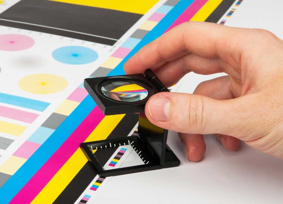 Eine Hand kontrolliert die Qualität eines farbigen CMYK-Druckbogens mit einem Fadenzähler.
