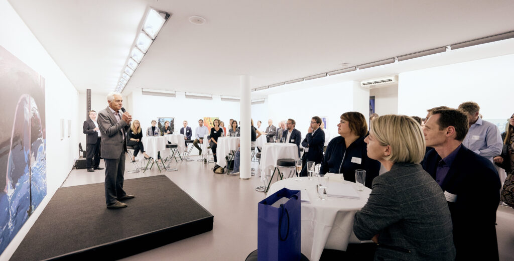 Ein Mann hält einen Vortrag mit Mikrofon vor Publikum in einer hellen Galerie.