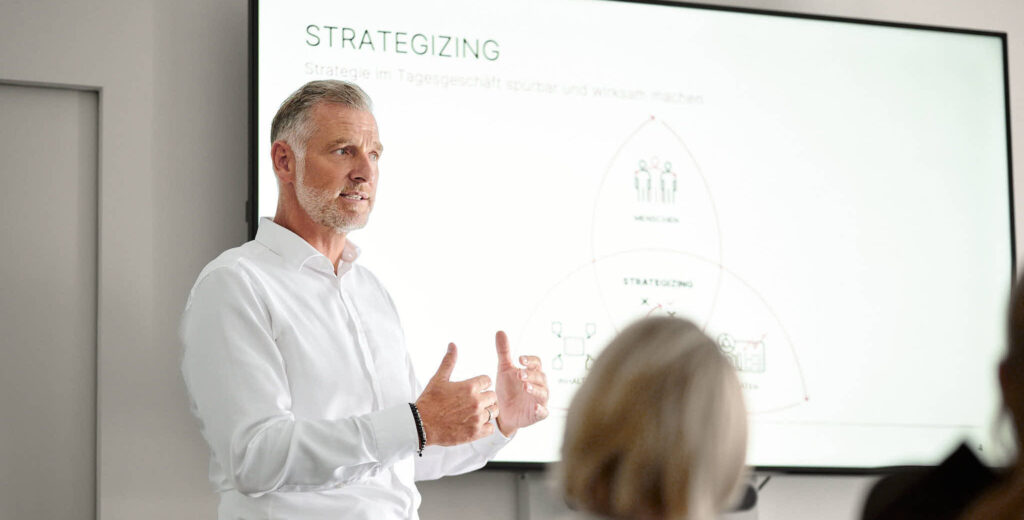 Ein Mann im weißen Hemd gestikuliert bei einer Präsentation zum Thema Strategie.