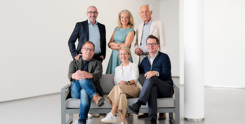 Sechs lächelnde Geschäftsleute posieren sitzend und stehend in einem hellen, modernen Raum.
