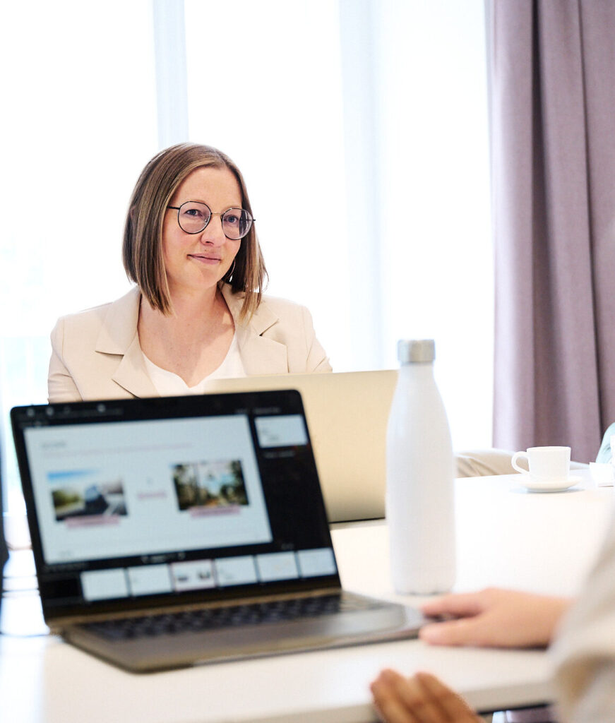 Eine lächelnde Frau mit Brille sitzt im Büro an einem Konferenztisch mit Laptops.