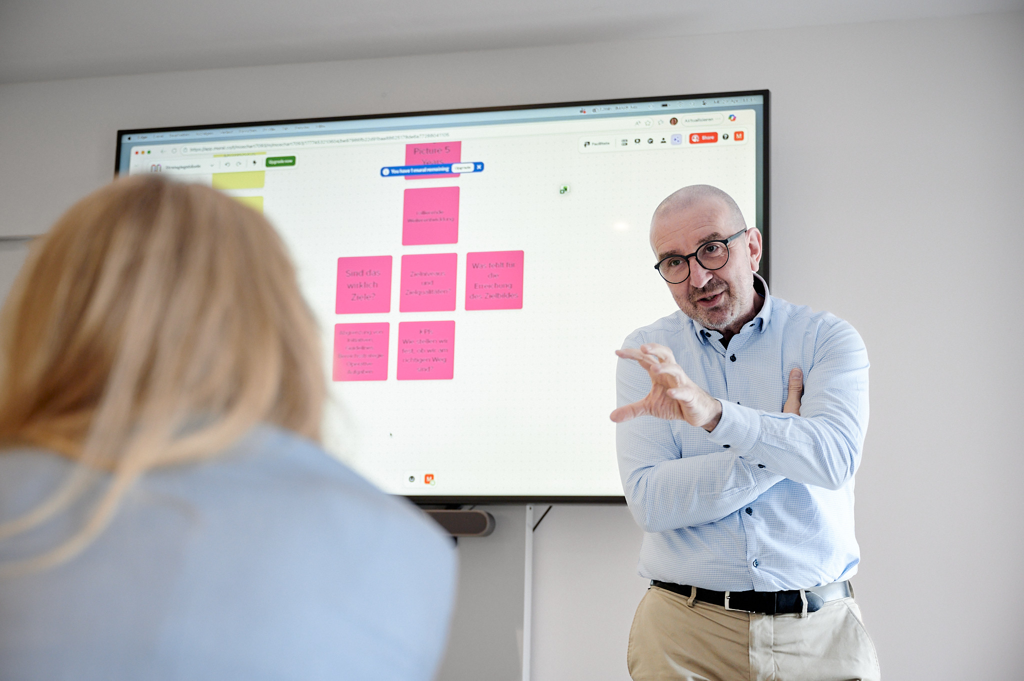 Ein Mann mit Brille gestikuliert vor einem Bildschirm mit rosa Haftnotizen während eines Workshops.