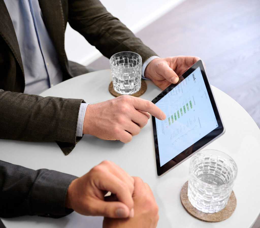 Ein Mann im Sakko zeigt seinem Gesprächspartner Daten auf einem Tablet.