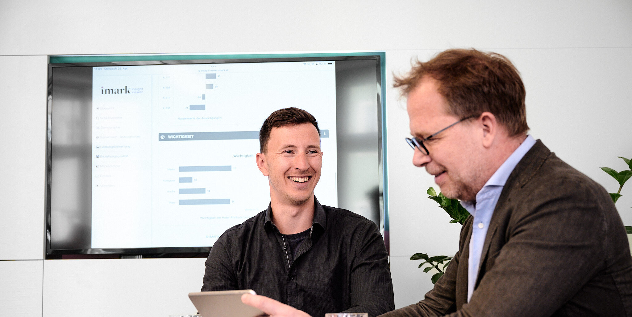 Zwei Männer besprechen lächelnd Diagramme vor einem großen Bildschirm im Büro.