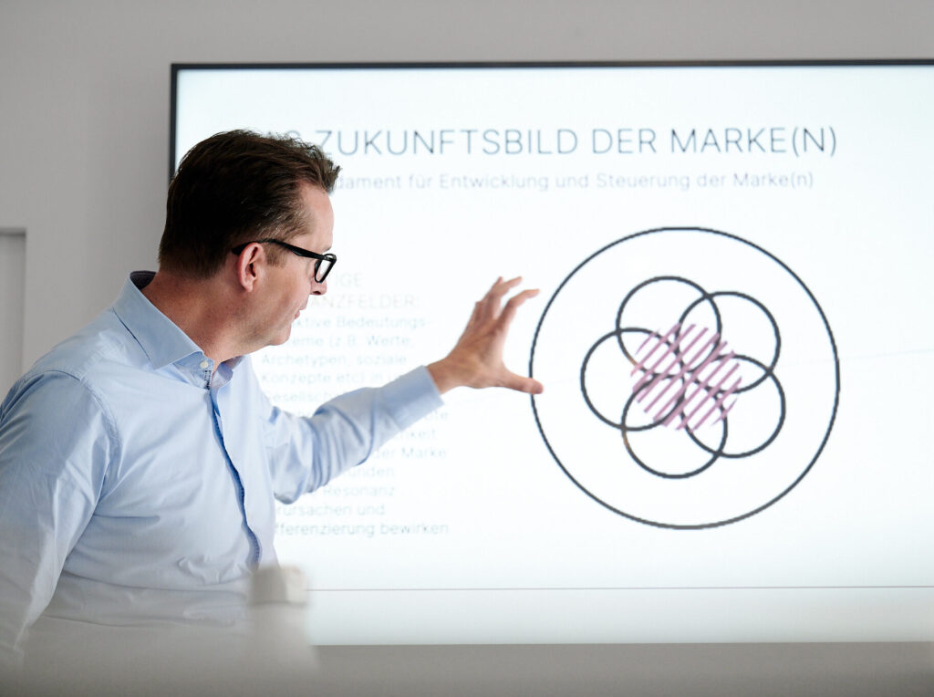 Ein Mann im hellblauen Hemd erklärt ein Diagramm auf einem großen Präsentationsbildschirm.