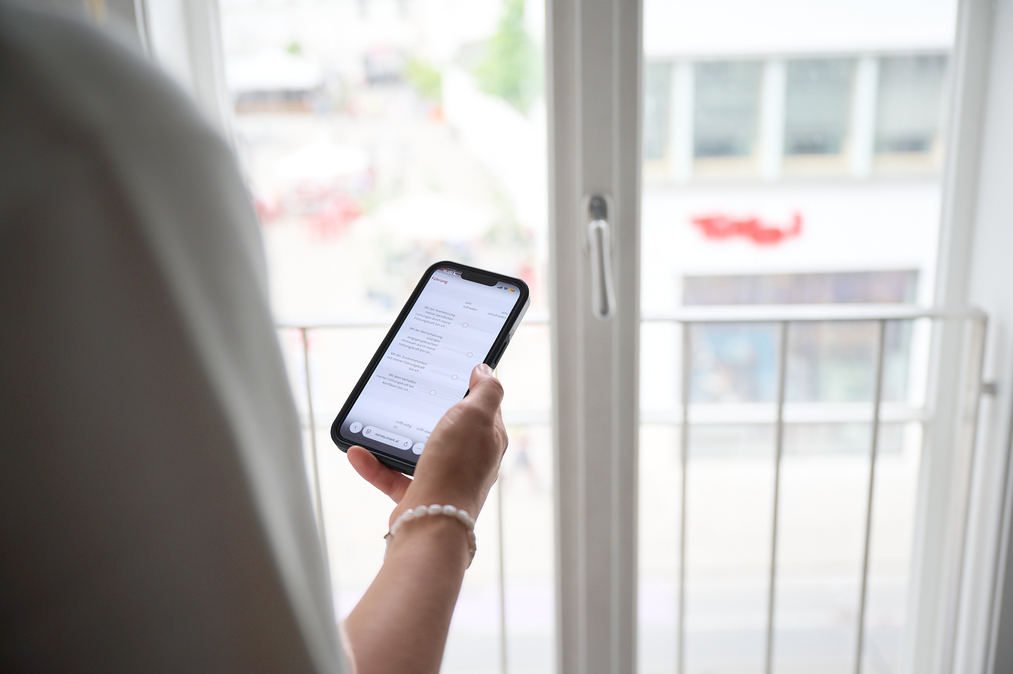 Eine Person hält ein Smartphone mit geöffneter App vor einem großen Balkonfenster.
