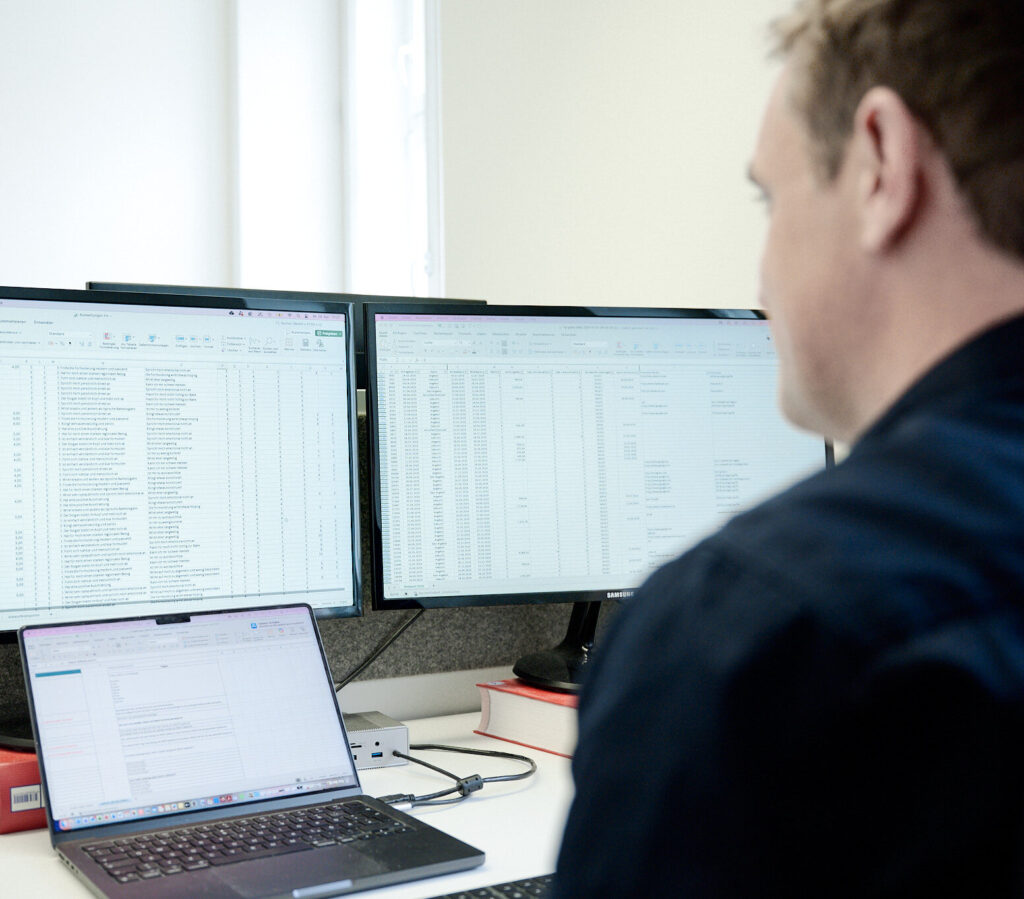 Ein Mitarbeiter analysiert Daten auf zwei großen Bildschirmen und einem Laptop am Arbeitsplatz.