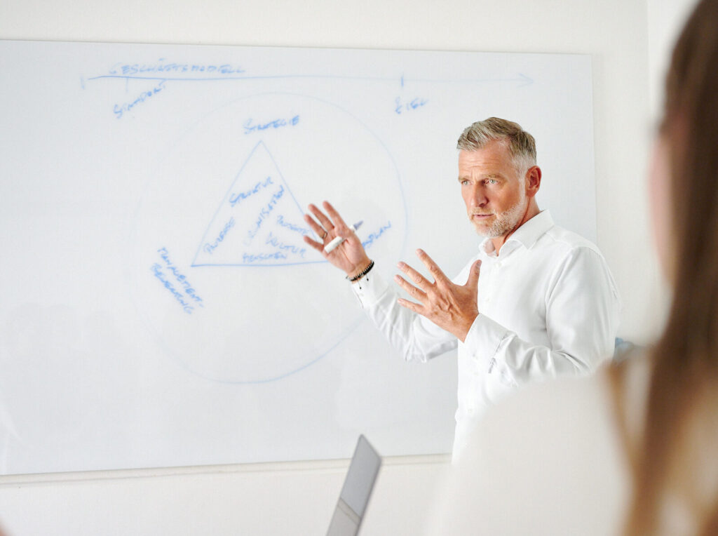 Ein ergrauter Mann erklärt gestikulierend eine Unternehmensstrategie an einem Whiteboard.