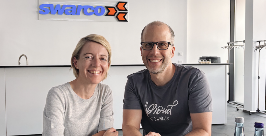 Eine blonde Frau und ein Mann mit Brille lächeln vor einem Swarco-Logo.