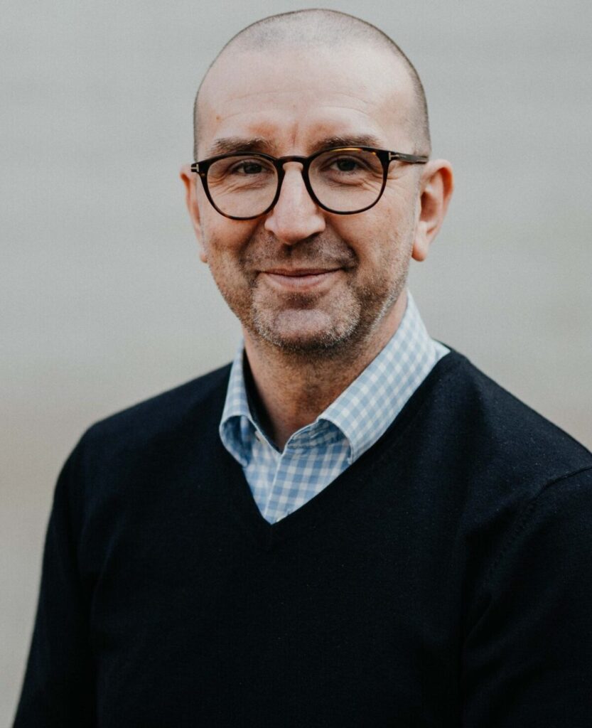 Lächelnder Mann mit Glatze, Brille, kariertem Hemd und schwarzem Pullover.