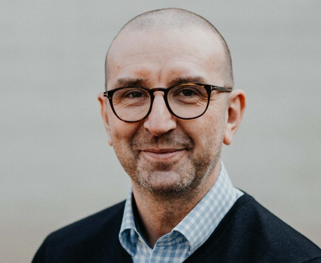 Lächelnder Mann mit Brille und Dreitagebart blickt freundlich direkt in die Kamera.