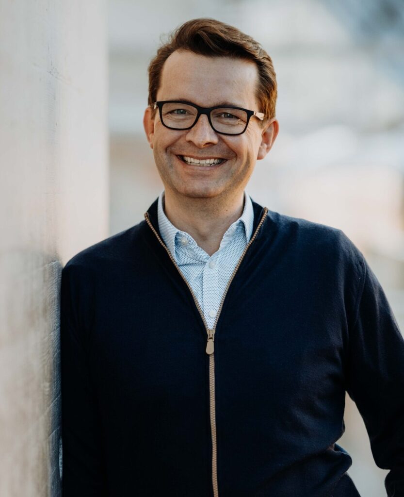 Lächelnder Mann mit Brille und dunkelblauer Strickjacke lehnt an einer hellen Wand.