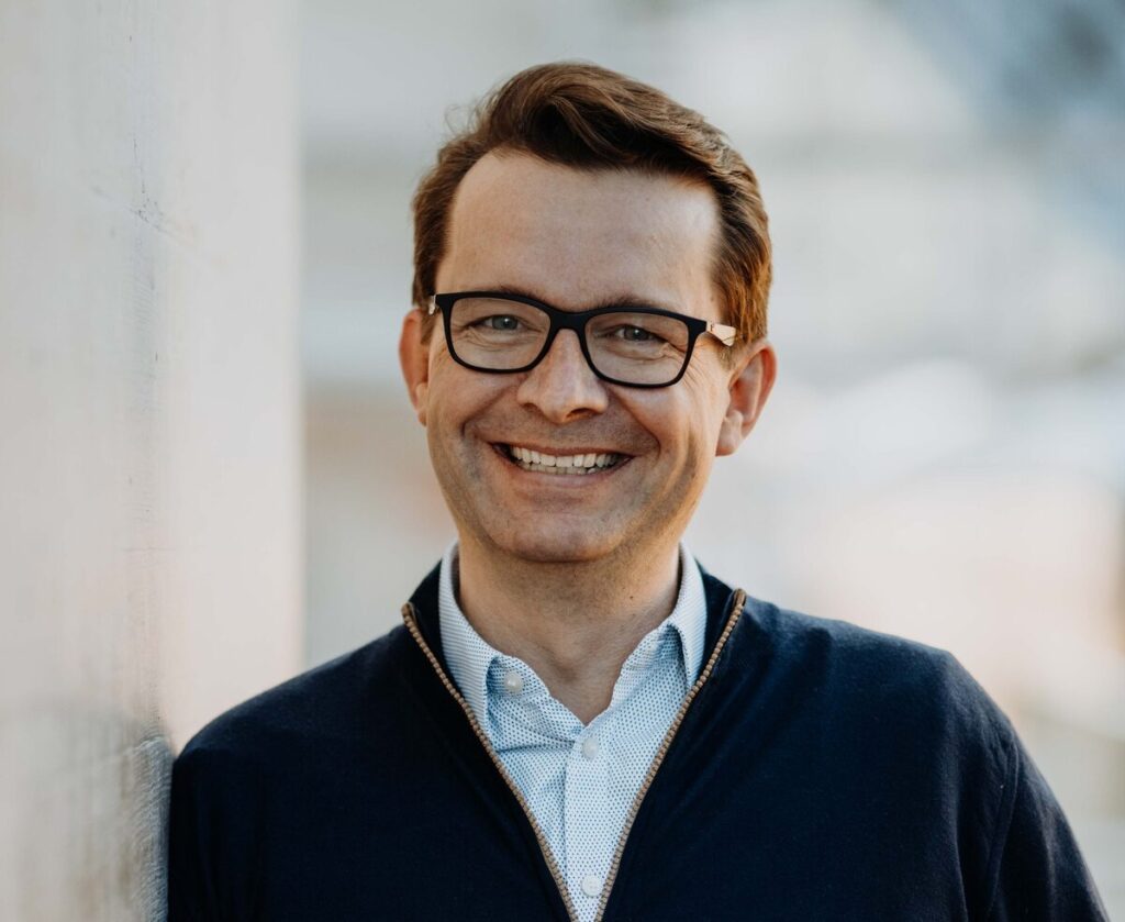 Lächelnder Mann mit Brille und dunkelblauem Pullover lehnt an einer Betonwand.