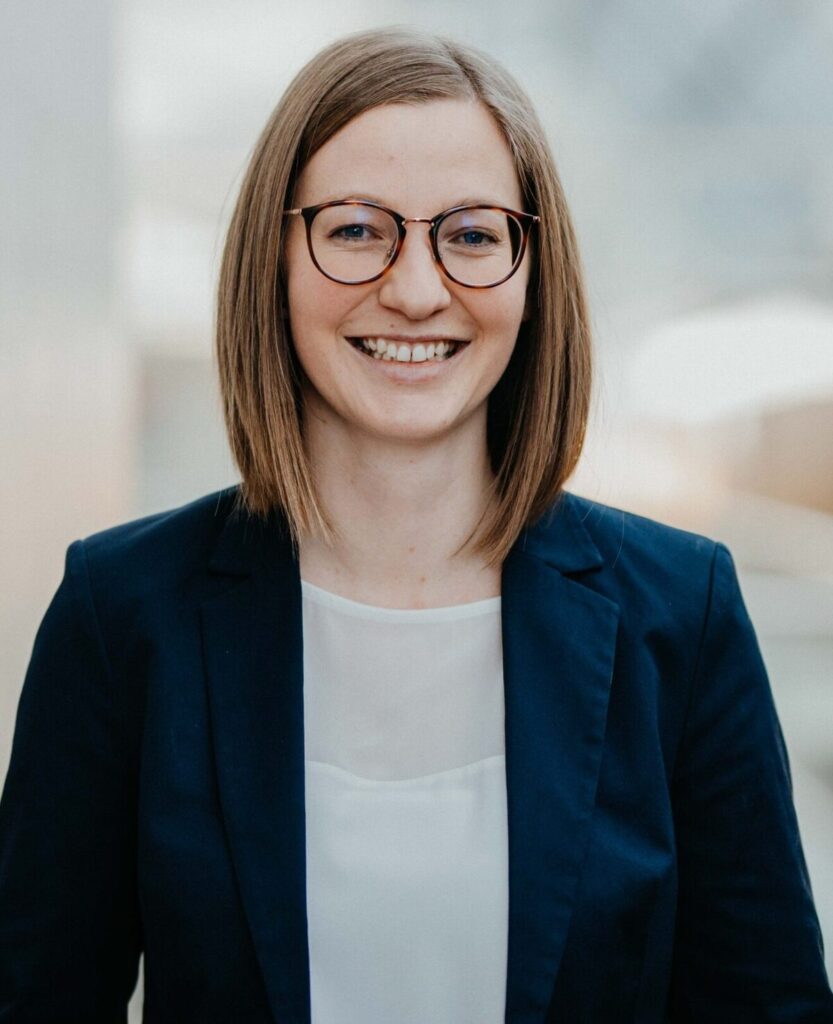 Eine lächelnde Frau mit Brille, kinnlangem Haar und dunkelblauem Blazer blickt in die Kamera.