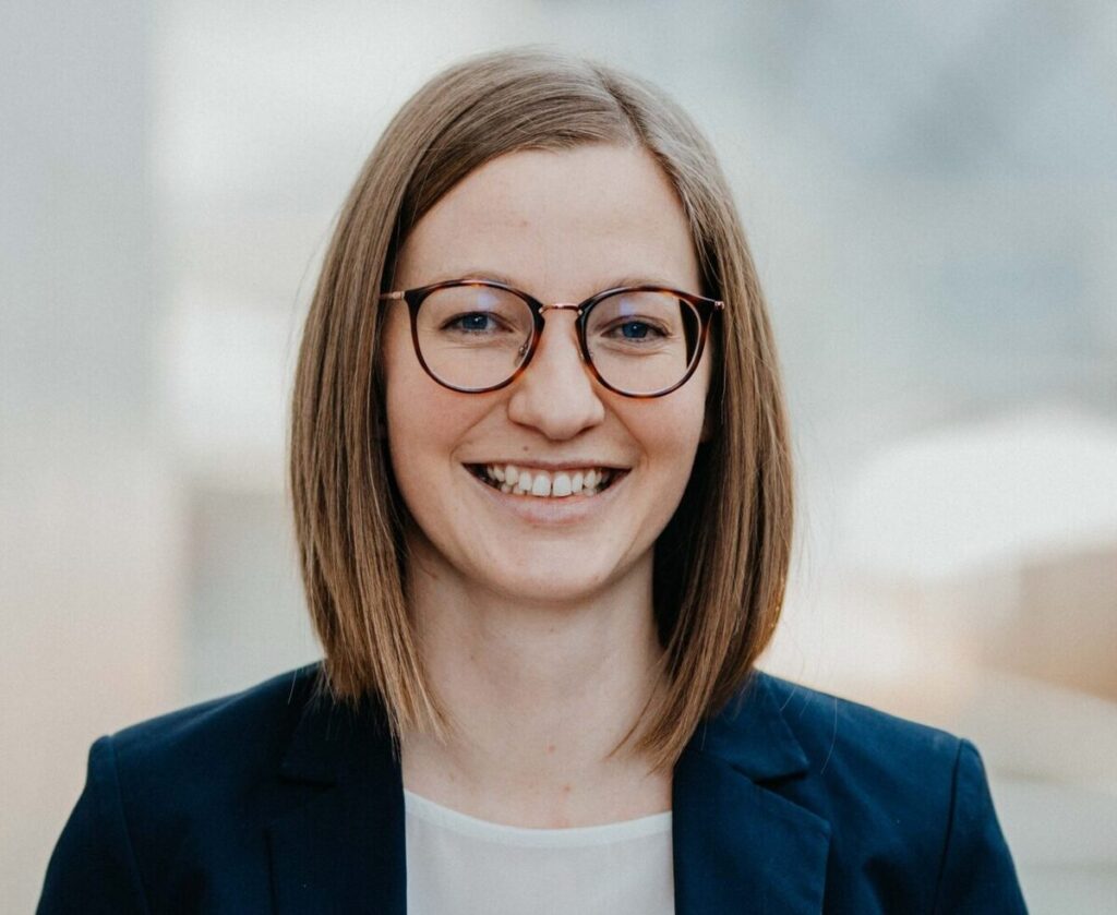 Eine lächelnde Frau mit Brille und dunkelblauem Blazer blickt direkt in die Kamera.