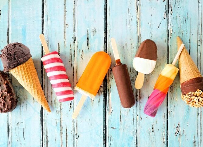 Verschiedene Eissorten am Stiel und in Waffeln liegen nebeneinander auf blauen Holzbrettern.