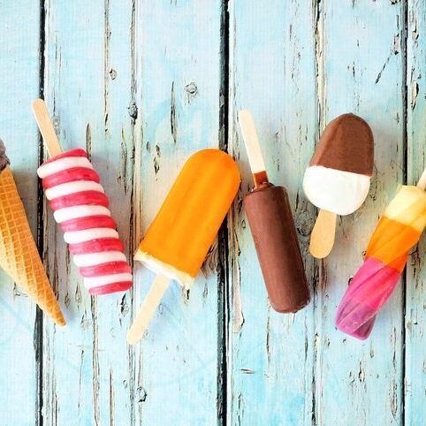 Verschiedene bunte Sorten Eis am Stiel liegen auf einem hellblauen Holzuntergrund.