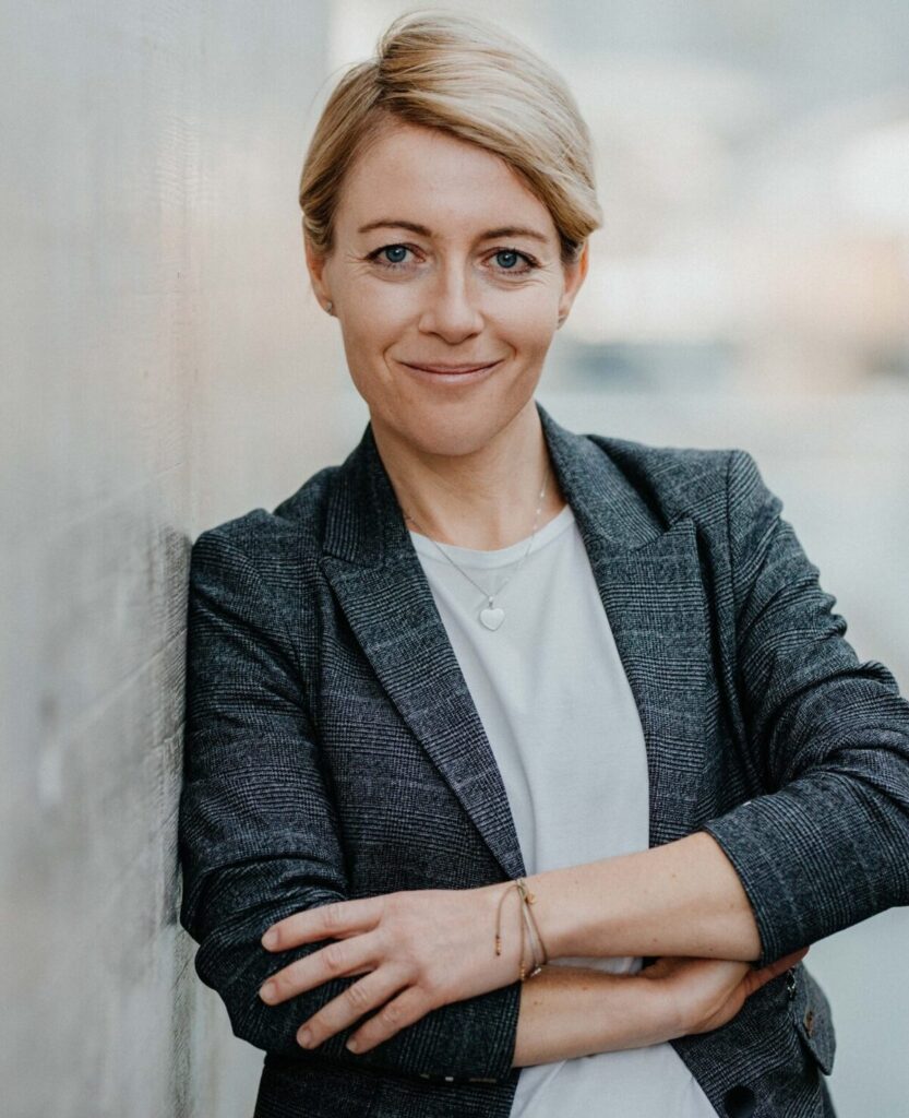 Lächelnde blonde Frau im grauen Blazer lehnt mit verschränkten Armen an einer Wand.