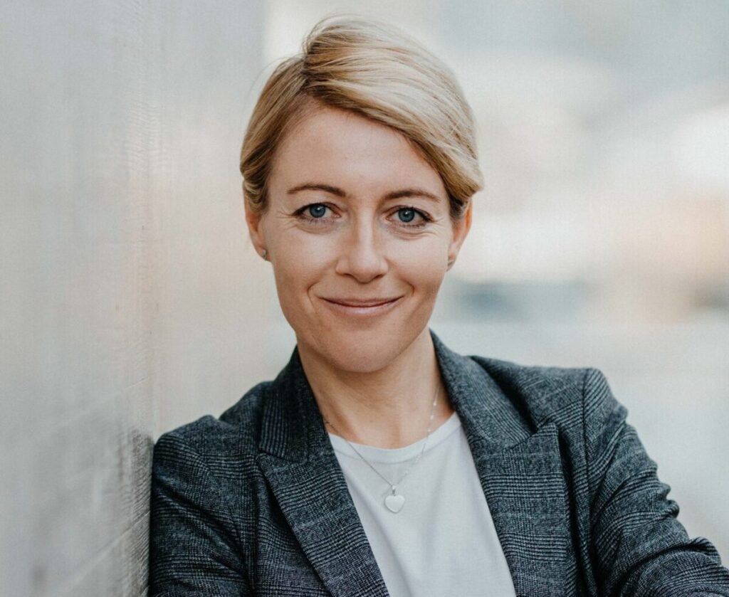 Blonde Frau im karierten grauen Blazer lächelt freundlich in die Kamera.
