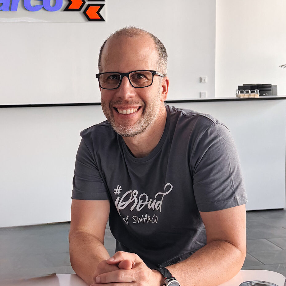 Ein lächelnder Mann mit Brille im grauen Swarco-Shirt sitzt an einem Tisch.