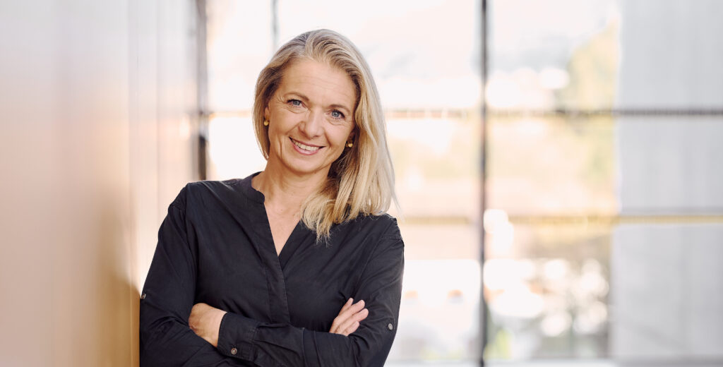 Eine lächelnde Frau mit blonden Haaren steht mit verschränkten Armen in einem modernen Büro.