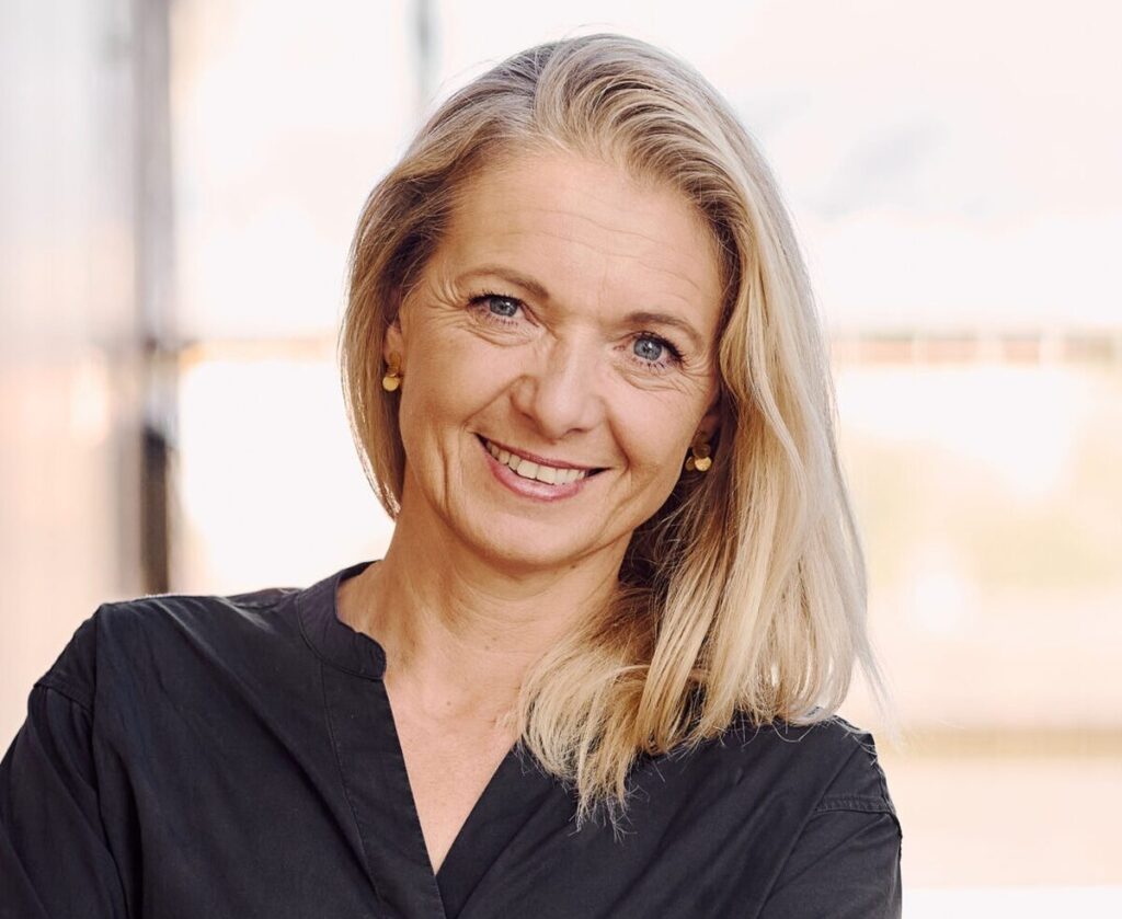 Eine freundlich lächelnde Frau mit blonden Haaren blickt direkt in die Kamera.