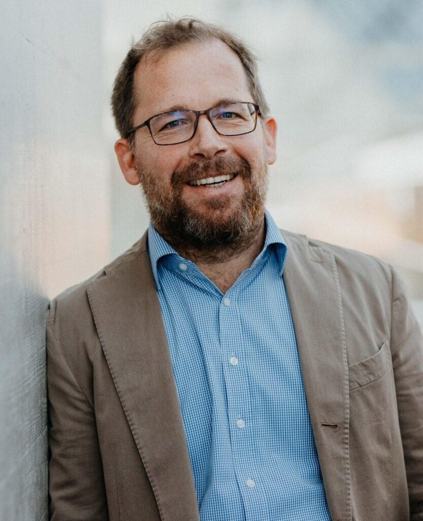 Lächelnder Mann mit Brille, Vollbart, blauem Hemd und braunem Sakko lehnt an einer Wand.