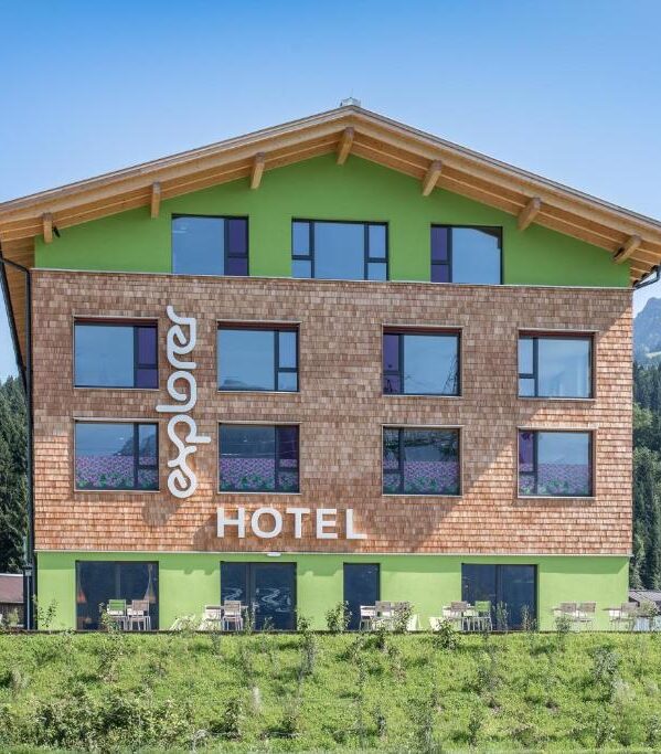 Außenansicht des Explorer Hotels mit Holzschindelfassade, grünen Akzenten und Sonnenterrasse.