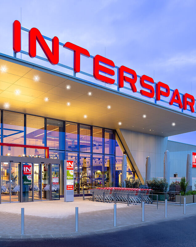 Moderne Interspar-Filiale mit beleuchtetem Eingang, rotem Schriftzug und Einkaufswagen in der Dämmerung.