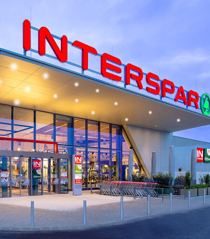Beleuchteter Eingang eines Interspar-Supermarktes mit aufgereihten Einkaufswagen im Abendlicht.