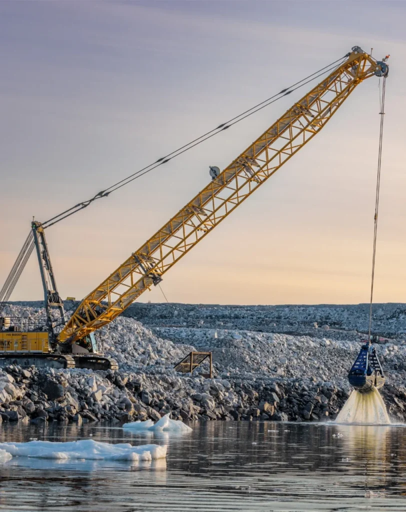 Ein gelber Gittermastkran baggert an einer felsigen Küste mit Eisschollen aus dem Wasser.