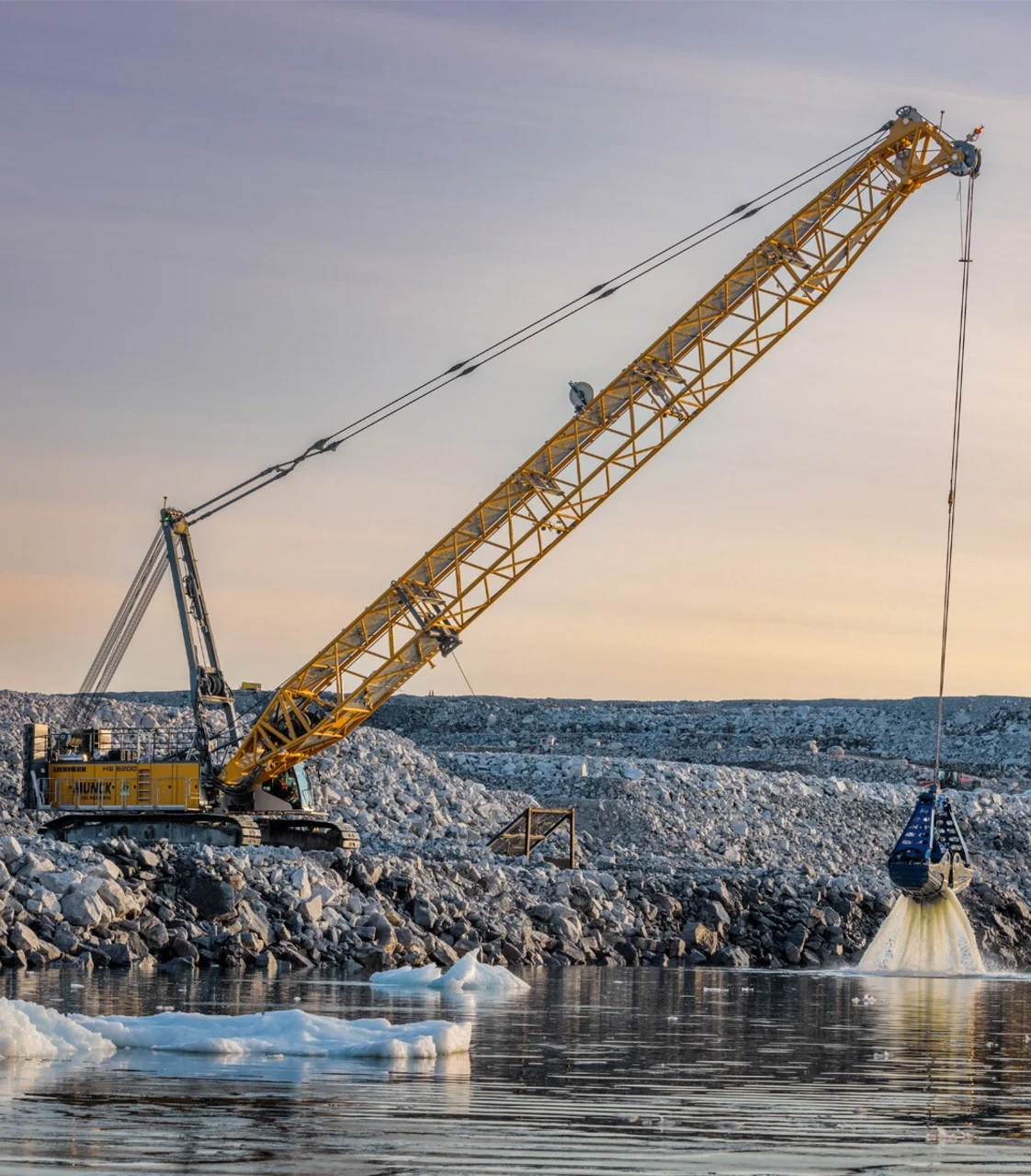 Ein gelber Gittermastkran baggert an einer felsigen Küste mit Eisschollen aus dem Wasser.