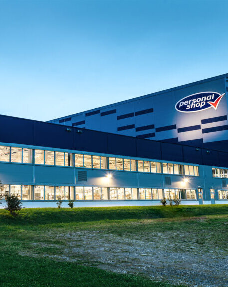 Modernes blaues Logistikzentrum der Marke Personalshop mit beleuchteten Fenstern bei klarem Himmel.