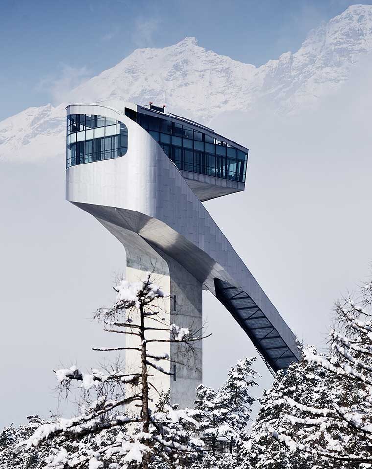 Futuristische Skisprungschanze mit verglastem Turm vor verschneiten Bergen und winterlichen Tannen.