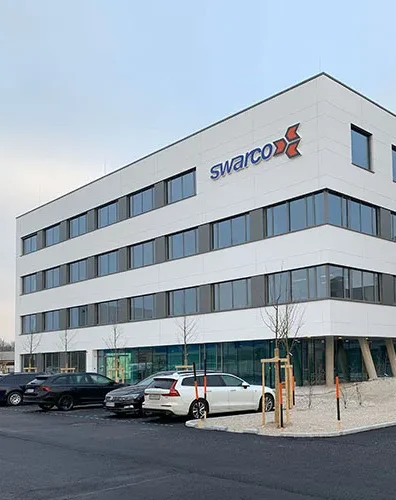 Modernes weißes Bürogebäude der Firma Swarco mit geparkten Autos auf dem Parkplatz.