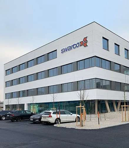 Modernes weißes Swarco-Bürogebäude mit Firmenlogo und geparkten Autos auf dem Parkplatz.